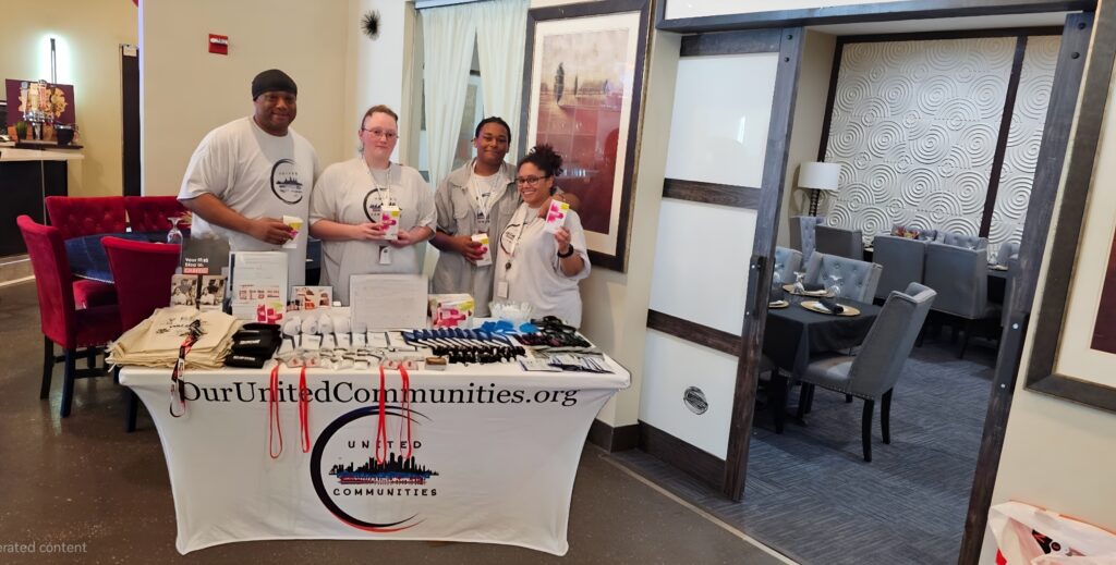 Four United Communities team members stand behind an outreach table displaying educational materials, Narcan kits, and branded items at a community event indoors. The table is covered with a United Communities logo tablecloth, and the group is smiling while holding outreach supplies.